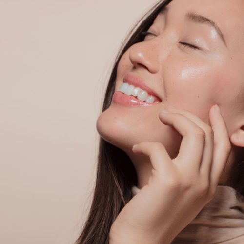 A woman with long dark hair smiles with her eyes closed, touching her cheek gently. Her skin looks smooth and radiant, and she is wearing a light-colored top against a plain beige background.