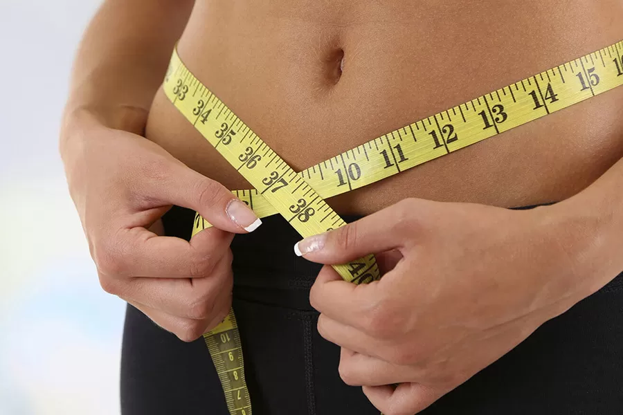 A person measures their waist with a yellow measuring tape, holding the tape around their bare midsection and wearing black pants.